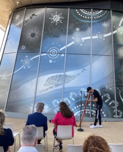 An Aboriginal man in black shirt and baseball cap plays a digeridoo into a microphone to an audience sat in white chairs in front of a giant mural on the windows of a building