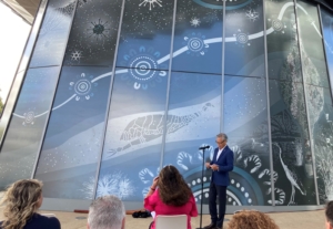 An Asian man a dark suit speaks into a microphone to an audience sat in white chairs in front of a giant mural on the windows of a building